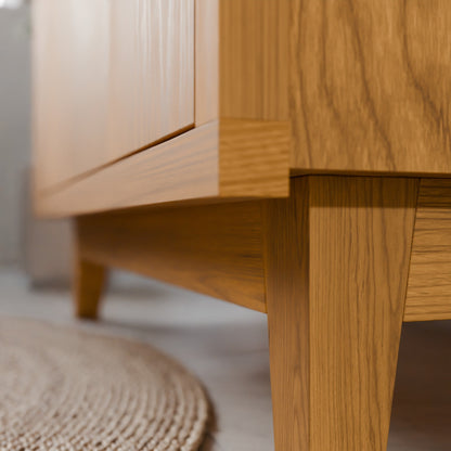 Close-up of a light oak wood furniture piece, showing the corner joint, leg, and cabinet door with a neutral background and a woven rug in the foreground.
