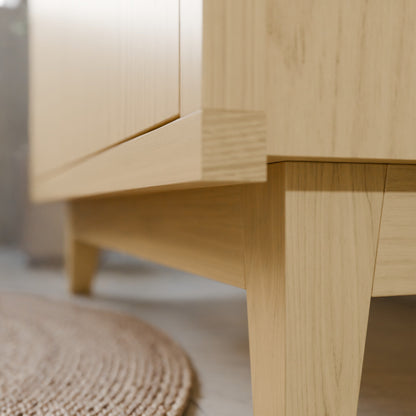 Close-up of a light natural wood furniture piece, showing the corner joint and leg detail against a softly blurred indoor background with a woven rug visible at the bottom left.