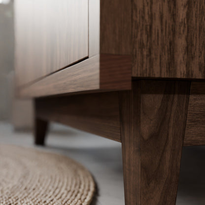 Close-up of a mid-century modern wooden cabinet or TV stand with a dark walnut finish, showing the grain and leg detail in a styled interior setting.