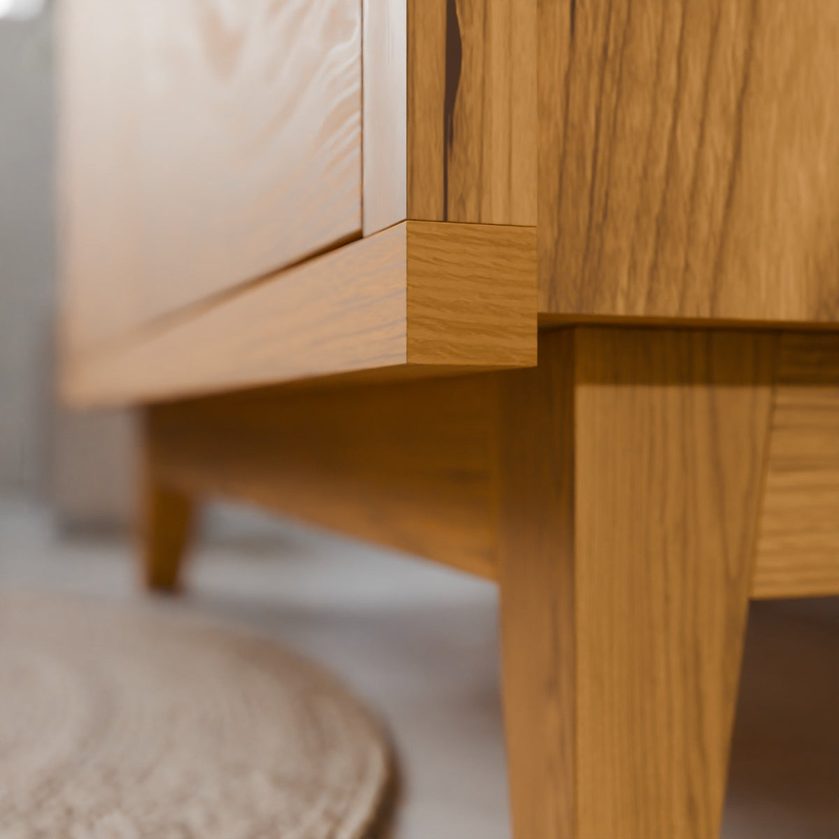Close-up detail of a light oak wood furniture piece, showing the corner joint, drawer front, and leg on a neutral indoor floor.