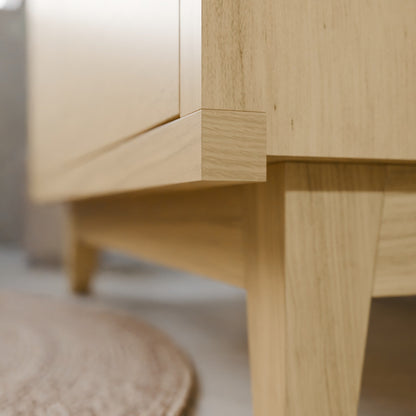 Close-up of a light natural wood furniture corner with visible grain, showing the joint between a cabinet door and leg in a softly blurred indoor setting.