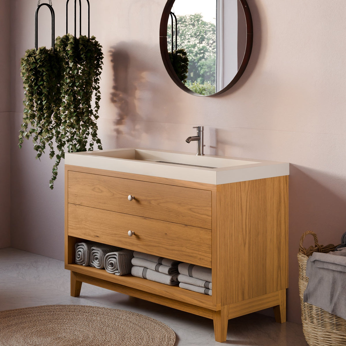A modern bathroom vanity cabinet with a light oak wood finish, featuring two drawers, a lower shelf with rolled towels, and a white integrated sink countertop. The cabinet is styled in a bathroom setting with a round mirror, hanging green plants, and a woven rug.