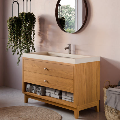 A modern bathroom vanity cabinet with a light oak wood finish, featuring two drawers, a lower shelf with rolled towels, and a white integrated sink countertop. The cabinet is styled in a bathroom setting with a round mirror, hanging green plants, and a woven rug.