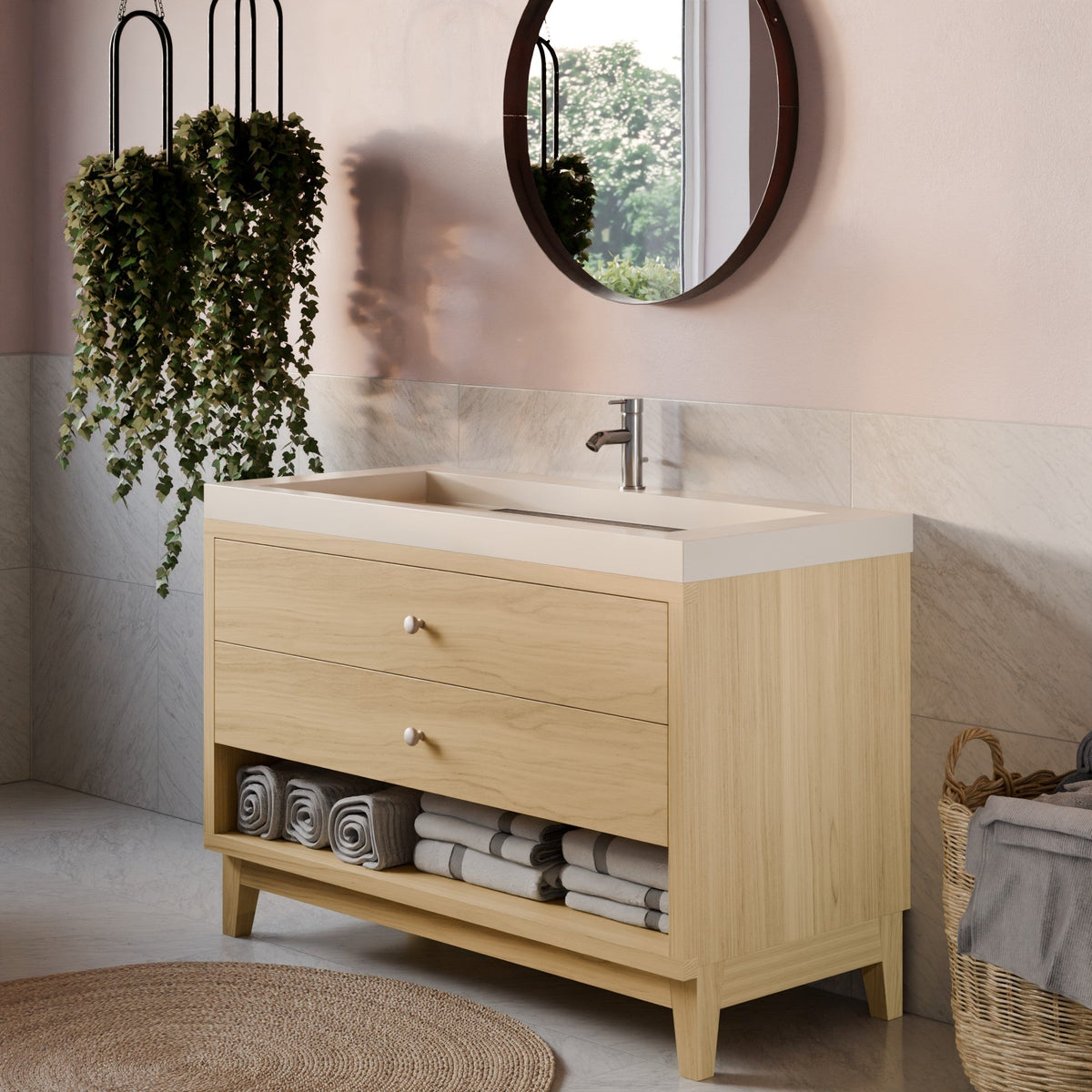 A light natural wood bathroom vanity with two drawers, an open lower shelf, and a white integrated sink basin with a chrome faucet. The vanity is styled in a bathroom setting with a round mirror, hanging green plants, rolled towels, and a woven rug.