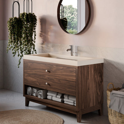 A modern bathroom vanity cabinet with a dark wood finish, a cream-colored stone sink top, and open lower storage shelves holding rolled towels. The vanity is styled in a bathroom setting with a round mirror, hanging green plants, a woven rug, and a basket.