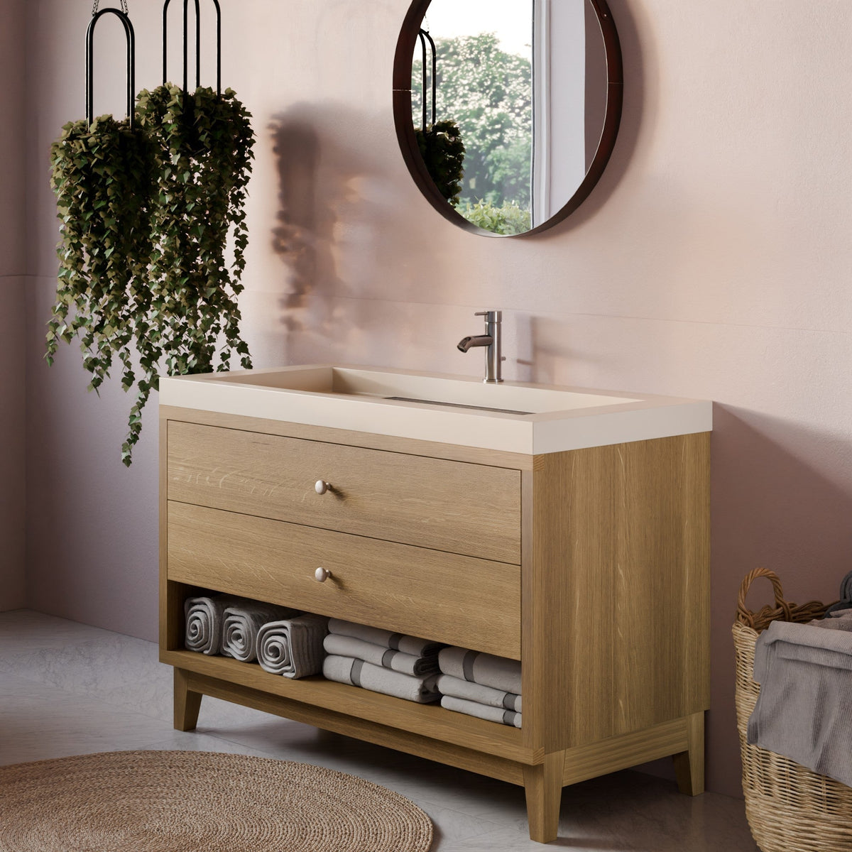 A light oak wood Krovel Furniture Co. bathroom vanity with two drawers, an open lower shelf, and a white integrated sink countertop, styled in a modern bathroom setting with a round mirror, hanging plants, and folded towels.