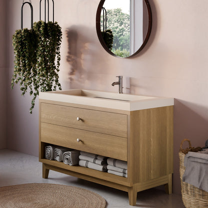 A light oak wood Krovel Furniture Co. bathroom vanity with two drawers, an open lower shelf, and a white integrated sink countertop, styled in a modern bathroom setting with a round mirror, hanging plants, and folded towels.