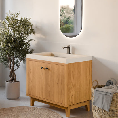 A modern bathroom vanity cabinet with a light oak wood finish and a white rectangular sink basin, styled in a bright bathroom next to a potted olive tree and a wicker basket.