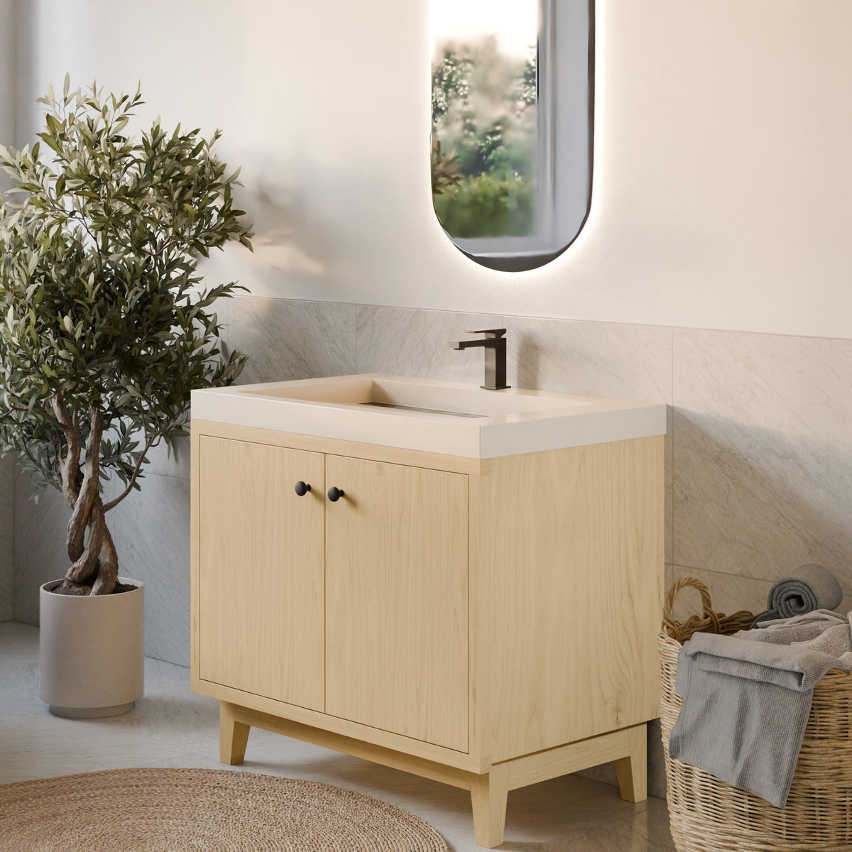 A light natural wood bathroom vanity cabinet with a white integrated sink countertop and a dark faucet, styled in a bright bathroom setting with a potted plant, woven rug, and basket.
