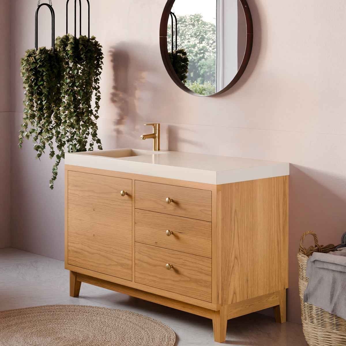 A light oak wood bathroom vanity with a white countertop and a gold faucet, styled in a modern bathroom setting with a pink wall, round mirror, hanging green plants, and a woven basket.