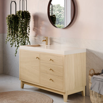 A light natural wood bathroom vanity with three drawers and a white integrated sink countertop, styled in a modern bathroom setting with a gold faucet, round mirror, hanging plants, and a woven rug.