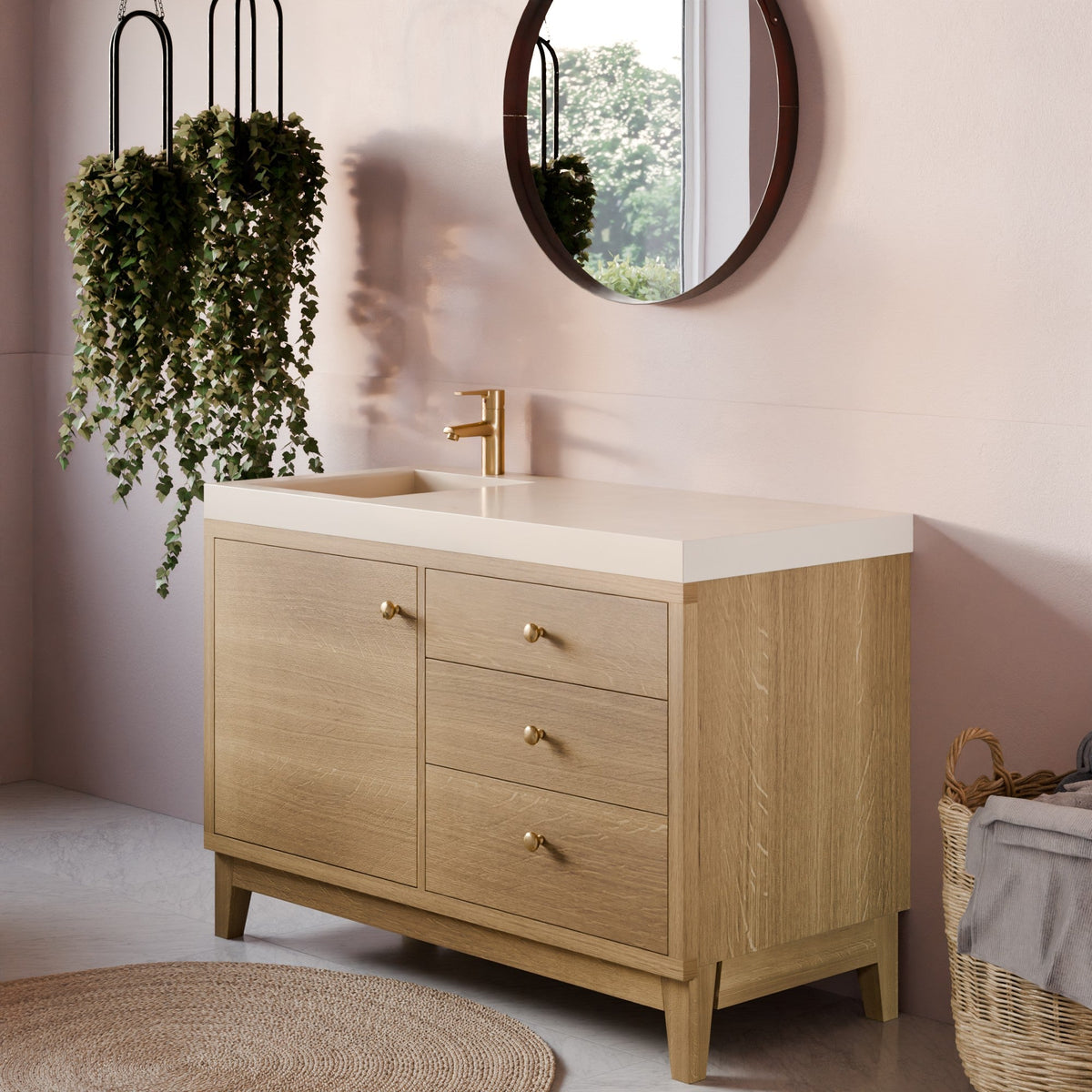 A light oak single-sink bathroom vanity with a white countertop and gold faucet, styled in a modern bathroom setting with a pink wall, round mirror, hanging greenery, and a woven basket.