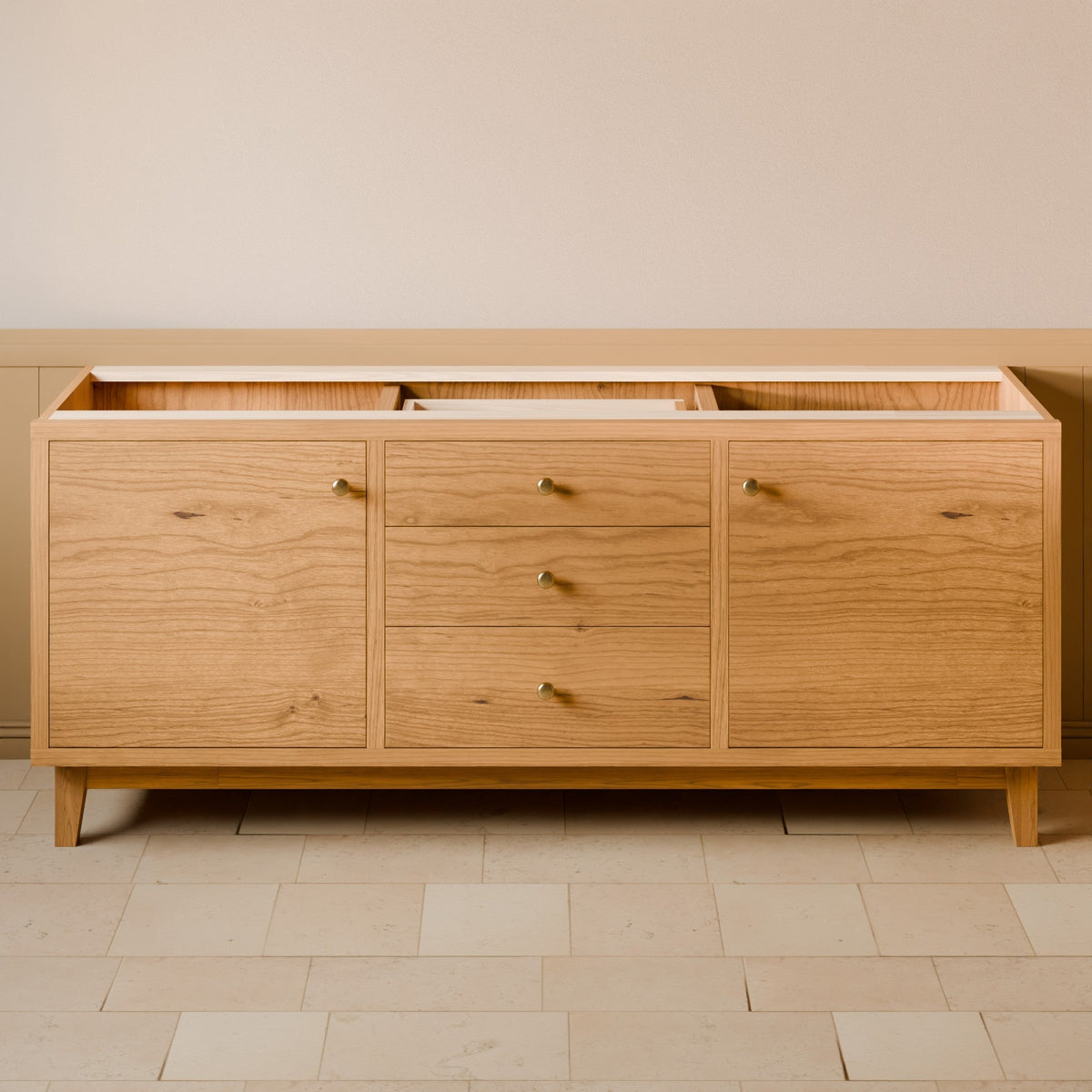 A mid-century modern oak wood sideboard cabinet with three drawers and three cabinet doors, featuring small brass knobs and tapered wooden legs, set against a neutral beige wall and tiled floor.