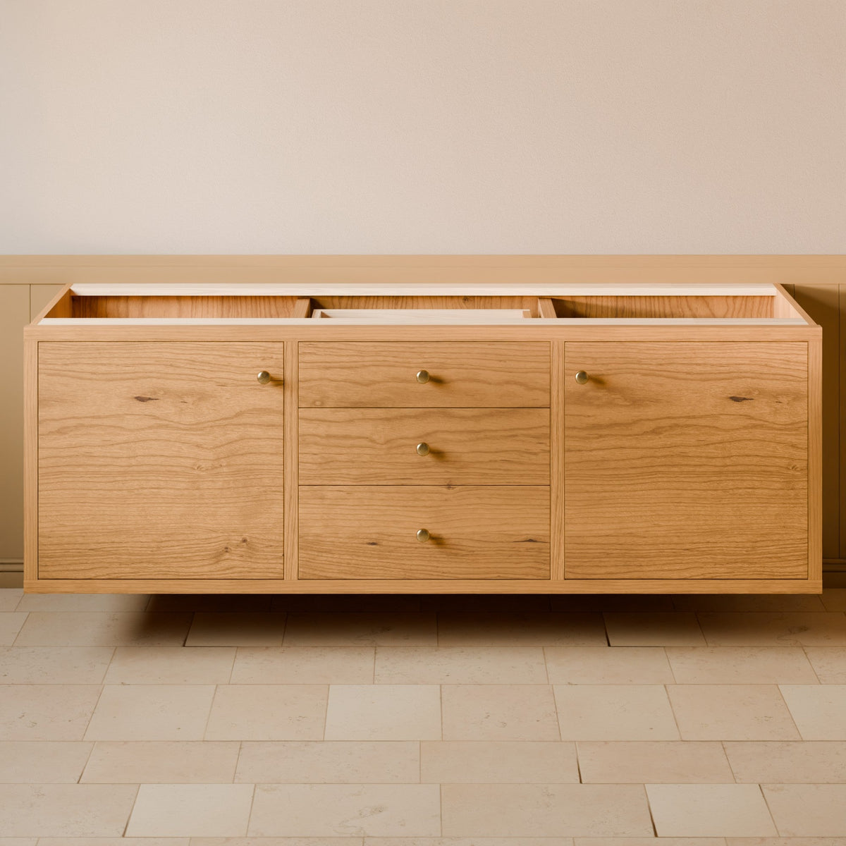 A light oak wood sideboard cabinet with three doors and three drawers, featuring small brass knobs, shown floating against a beige wall and light stone tile floor.
