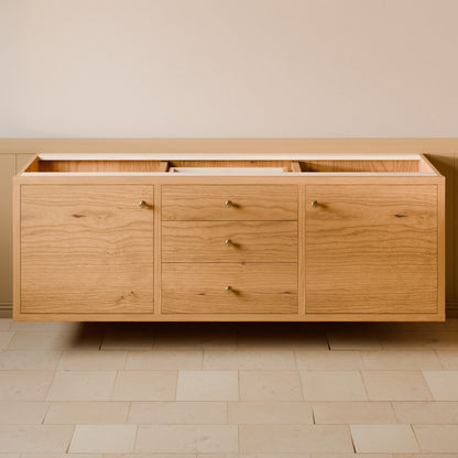 A light oak wood sideboard cabinet with three doors and three drawers, featuring small brass knobs, shown floating against a beige wall and light stone tile floor.