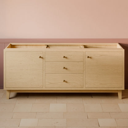 A light natural oak wood sideboard or credenza with three drawers in the center and two cabinet doors on the sides, accented with small round brass knobs, set against a pink and beige wall with tiled flooring.