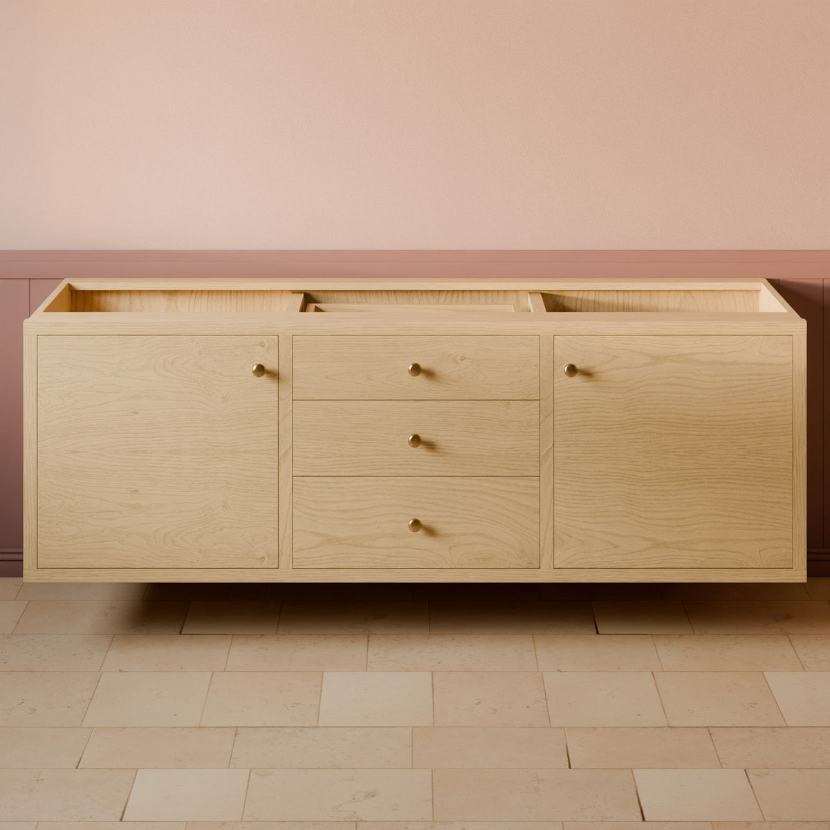 A floating light oak wood bathroom vanity cabinet with three drawers and two side doors, featuring small brass round knobs, set against a pink and brown wainscoted wall and tiled floor.