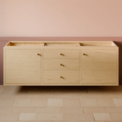 A floating light oak wood bathroom vanity cabinet with three drawers and two side doors, featuring small brass round knobs, set against a pink and brown wainscoted wall and tiled floor.