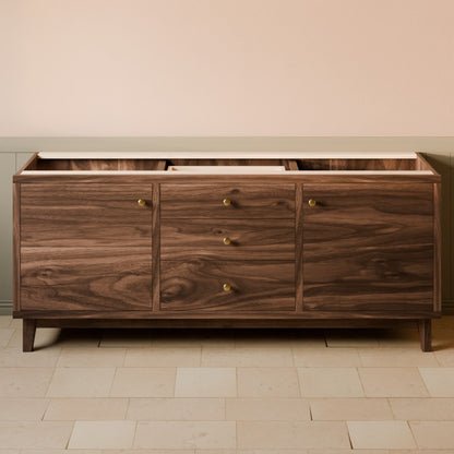 A mid-century modern wooden sideboard or credenza with a dark walnut finish, featuring three drawers with brass knobs and an open top shelf, set against a beige wall and light stone tile floor.