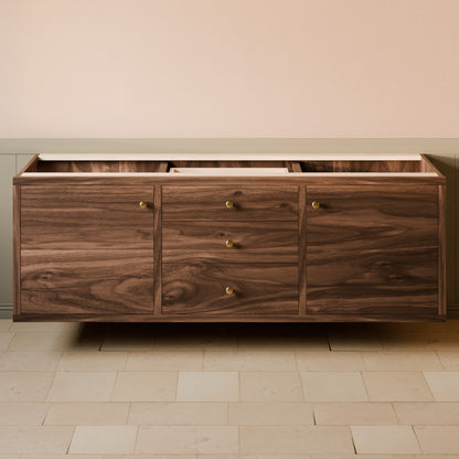 A floating wooden sideboard or credenza with three drawers and three cabinet doors, featuring a dark walnut wood grain finish and small brass knobs, set against a beige wall and light stone tile floor.