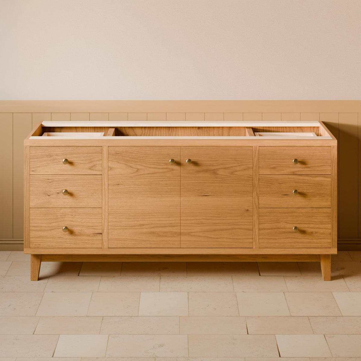 A light oak wood sideboard or credenza with six drawers and two cabinet doors, featuring small brass handles and tapered legs, set against a beige wall with wainscoting and light stone tile flooring.