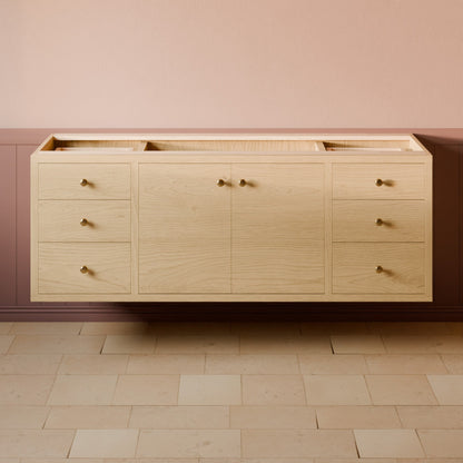 A light natural wood bathroom vanity cabinet with multiple drawers and two central doors, mounted against a pink wall above a beige stone floor.