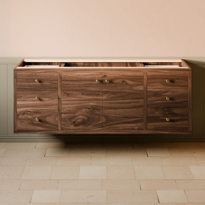 A floating wooden credenza or sideboard with a rich walnut wood grain finish, featuring six drawers with brass knobs, mounted against a beige wall above a light stone tile floor.