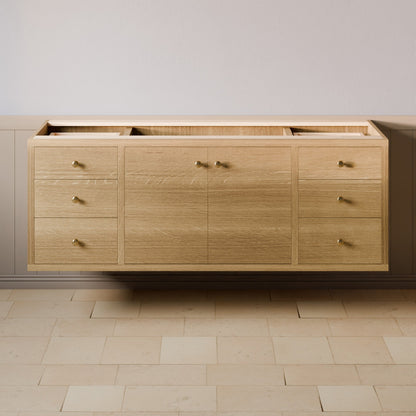 A light oak wood wall-mounted vanity cabinet with multiple drawers and doors, featuring small brass handles, shown against a neutral beige wall and tiled floor.