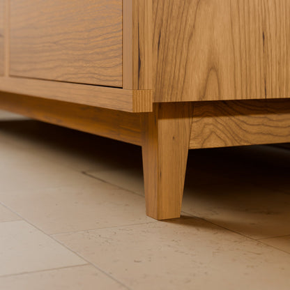 Close-up of a mid-century modern wooden cabinet with a warm oak finish, showing the corner joint and tapered legs on a light beige tiled floor.