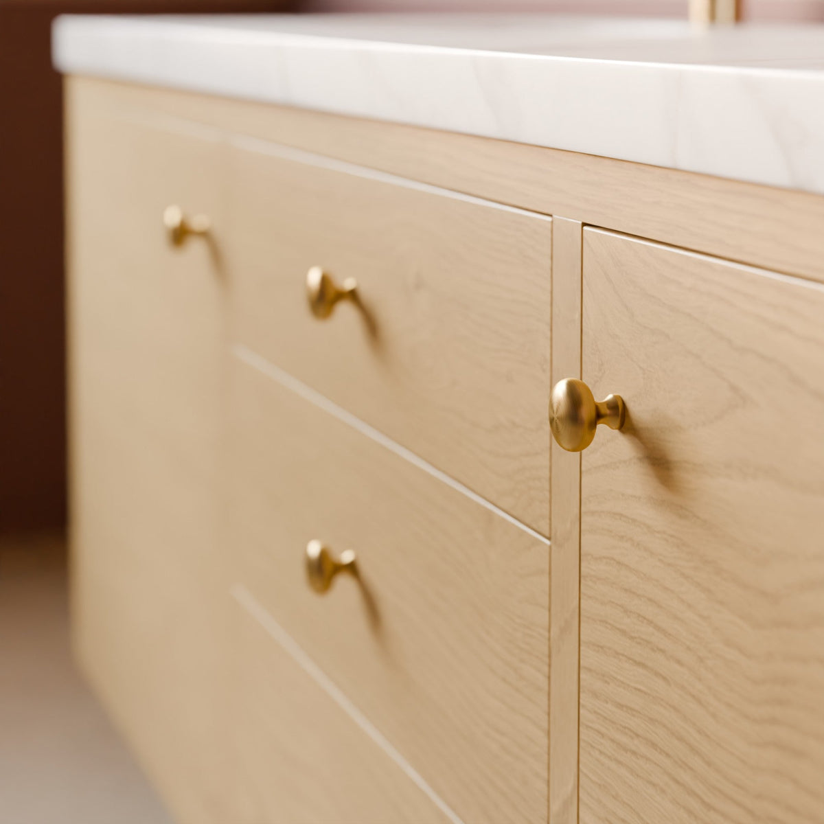 Close-up of a light wood vanity cabinet with three drawers and one door, each fitted with round brass knobs, topped with a white marble countertop.