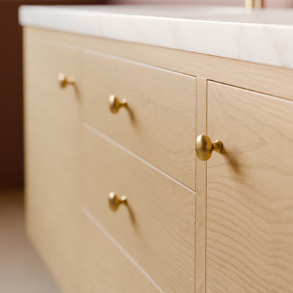 Close-up of a light wood vanity cabinet with three drawers and one door, each fitted with round brass knobs, topped with a white marble countertop.