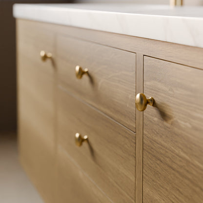 Close-up of a light oak wood vanity cabinet with three drawers and one cabinet door, accented with round brass knobs and topped with a white marble countertop.