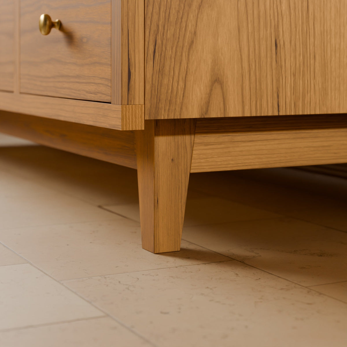 Close-up of a light oak wood cabinet or sideboard showing the corner joint, drawer front with a brass knob, and front leg on a beige tiled floor.