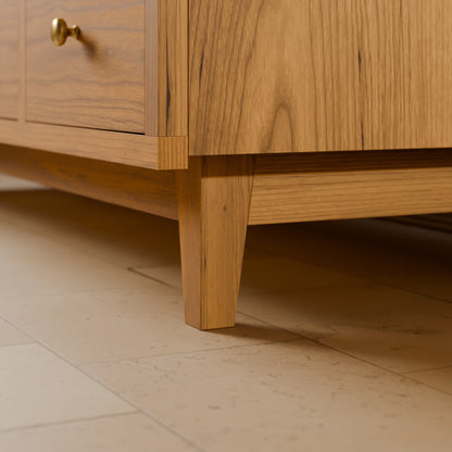 Close-up of a light oak wood cabinet or sideboard showing the corner joint, drawer front with a brass knob, and front leg on a beige tiled floor.