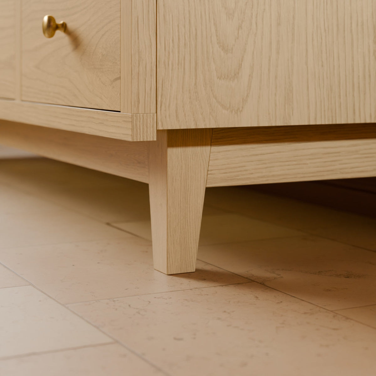 Close-up of a light natural oak wooden cabinet or dresser leg and drawer corner, with a small brass knob visible, set against a matching stone tile floor.