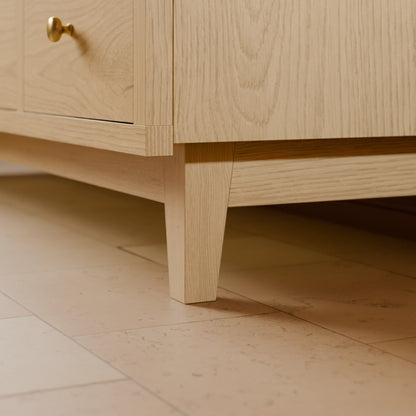 Close-up of a light natural oak wooden cabinet or dresser leg and drawer corner, with a small brass knob visible, set against a matching stone tile floor.