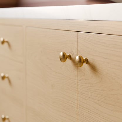 Close-up of a light natural wood cabinet with two round brass knobs and a white countertop edge.