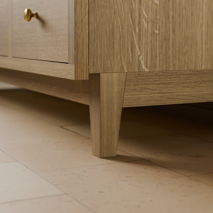 Close-up of a light oak wood cabinet or sideboard showing a drawer with a brass knob, the cabinet leg, and the tiled floor below.