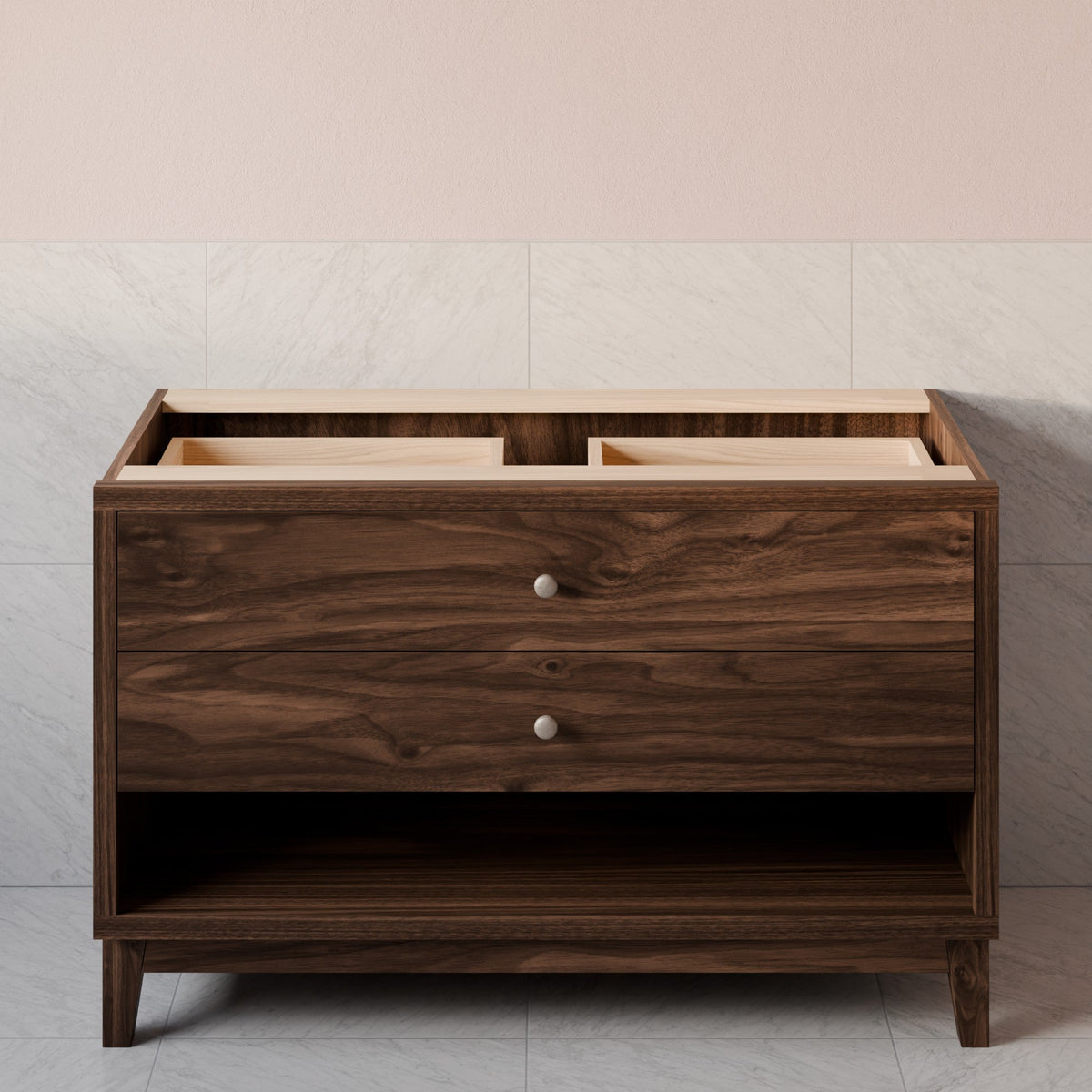 A dark brown wooden bathroom vanity cabinet with two drawers, round silver knobs, and an open lower shelf, set against a light gray marble-tiled wall and pale pink upper wall.