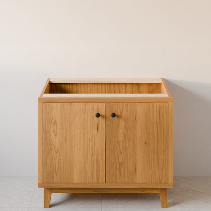 A light oak wood cabinet with two doors, small black round knobs, and a built-in open top shelf, set against a plain off-white wall.