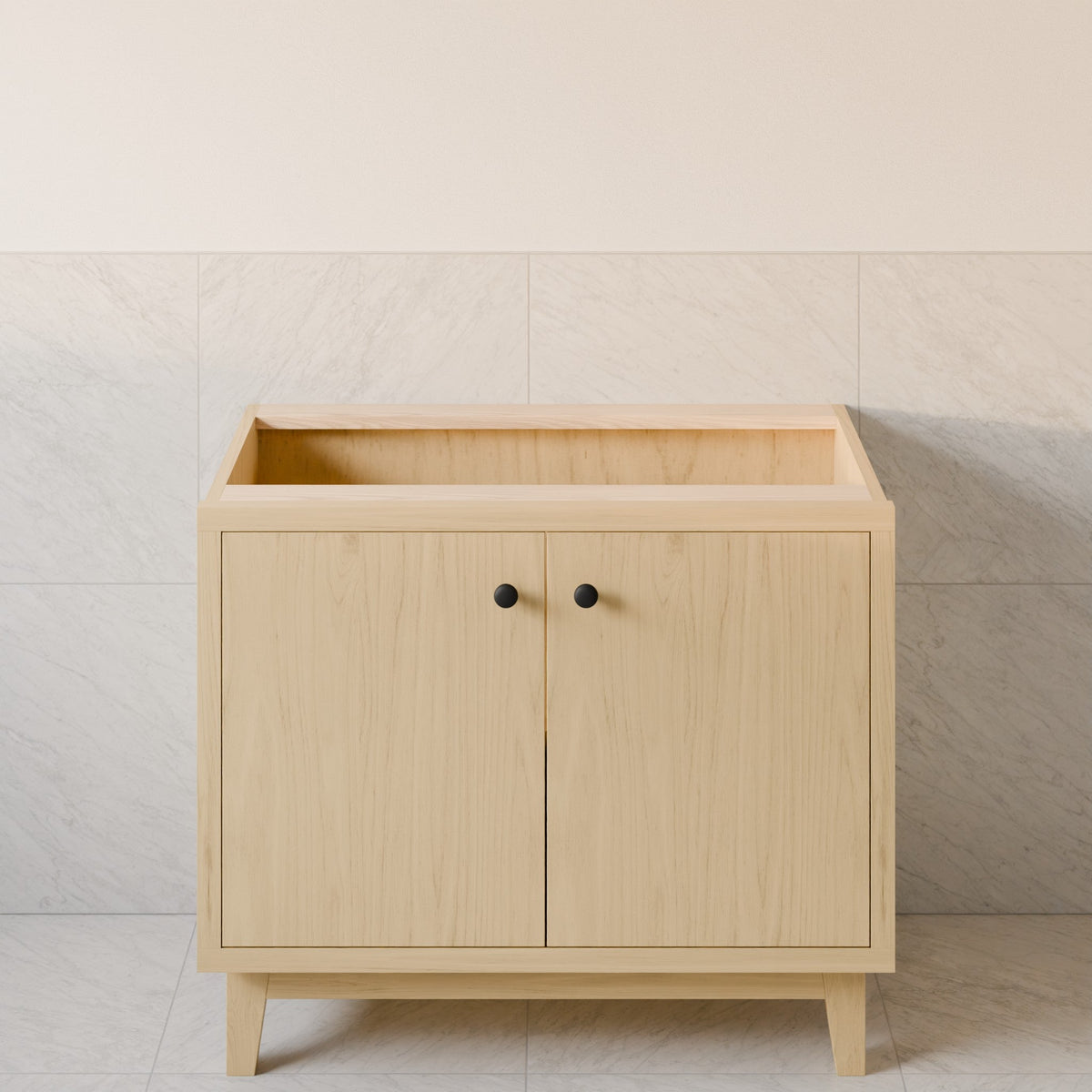 A light natural wood bathroom vanity cabinet with two doors, small round black knobs, and tapered legs, set against a tiled wall.