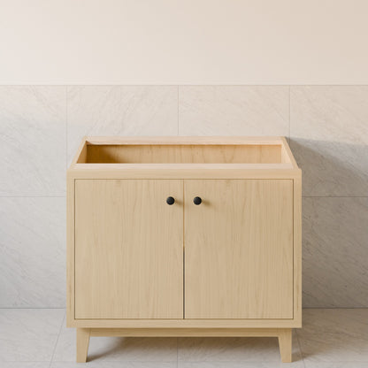 A light natural wood bathroom vanity cabinet with two doors, small round black knobs, and tapered legs, set against a tiled wall.