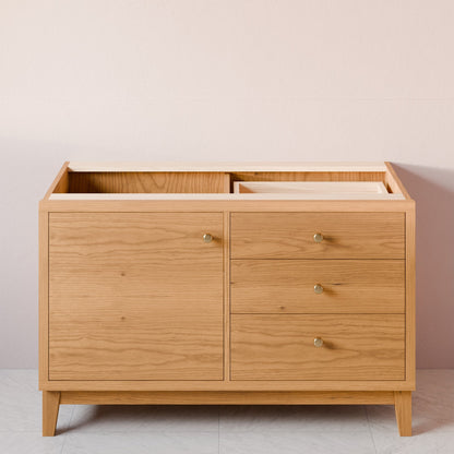 A light oak wood changing table with a top storage compartment, three drawers on the right with small round brass pulls, and two open doors on the left, shown against a neutral beige wall.