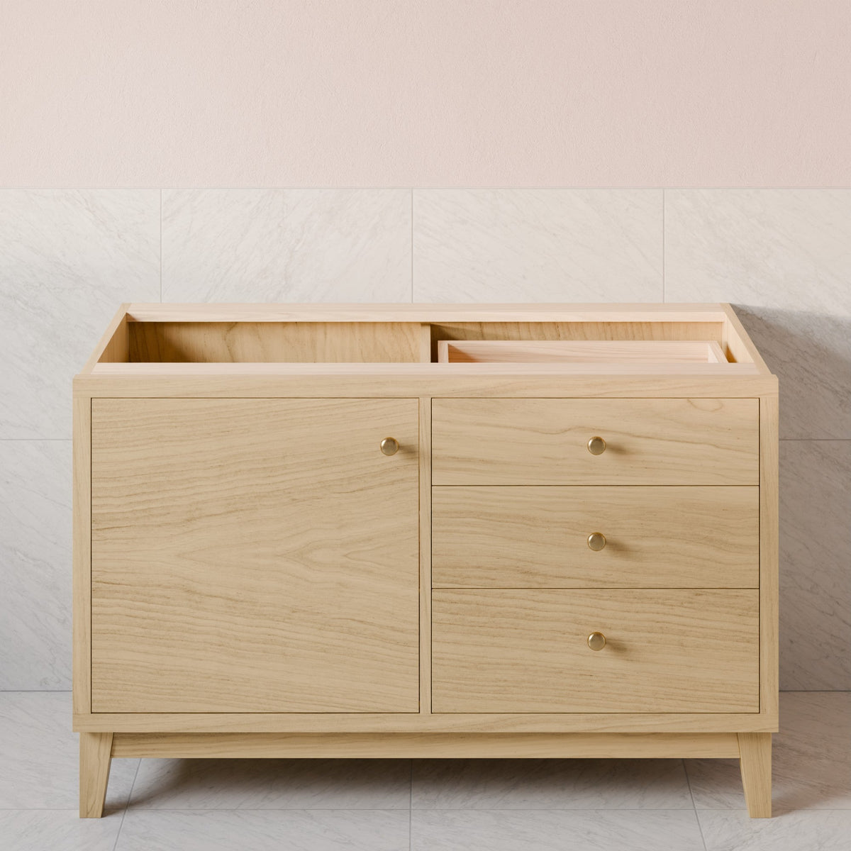 A light natural wood bathroom vanity cabinet with a single door and three drawers, featuring small round brass knobs and tapered legs, set against a pale pink and white tiled wall.