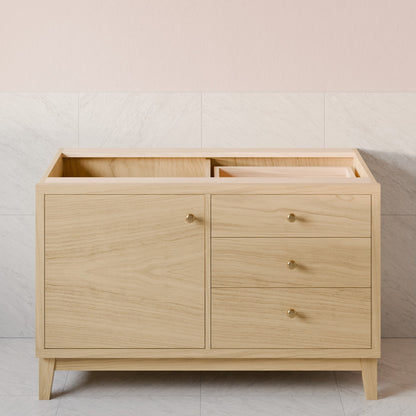 A light natural wood bathroom vanity cabinet with a single door and three drawers, featuring small round brass knobs and tapered legs, set against a pale pink and white tiled wall.