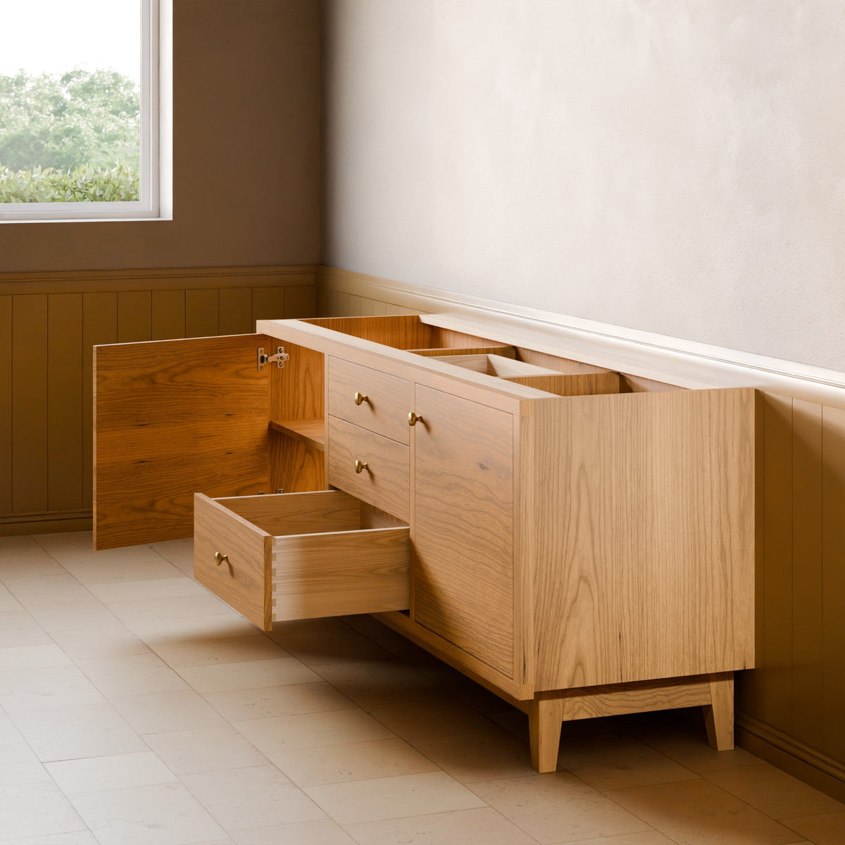 A mid-century modern light oak wood sideboard or credenza with brass handles, shown in a room setting with one cabinet door and one drawer pulled open. The room features a textured off-white wall, brown wainscoting, and light-colored tile flooring.