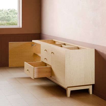 A light natural wood storage cabinet with multiple drawers and an open top compartment, shown in a styled room corner with brown wainscoting, a beige wall, and a window.