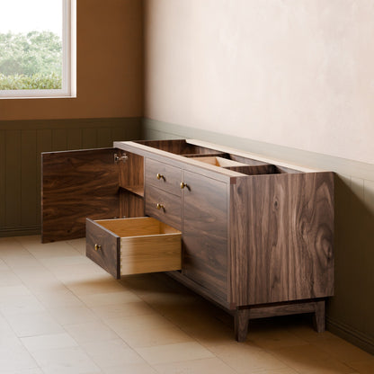 A dark wood sideboard or credenza with a rich walnut-like grain, featuring brass handles and an open lower drawer, set in a styled room with beige walls and tiled flooring.