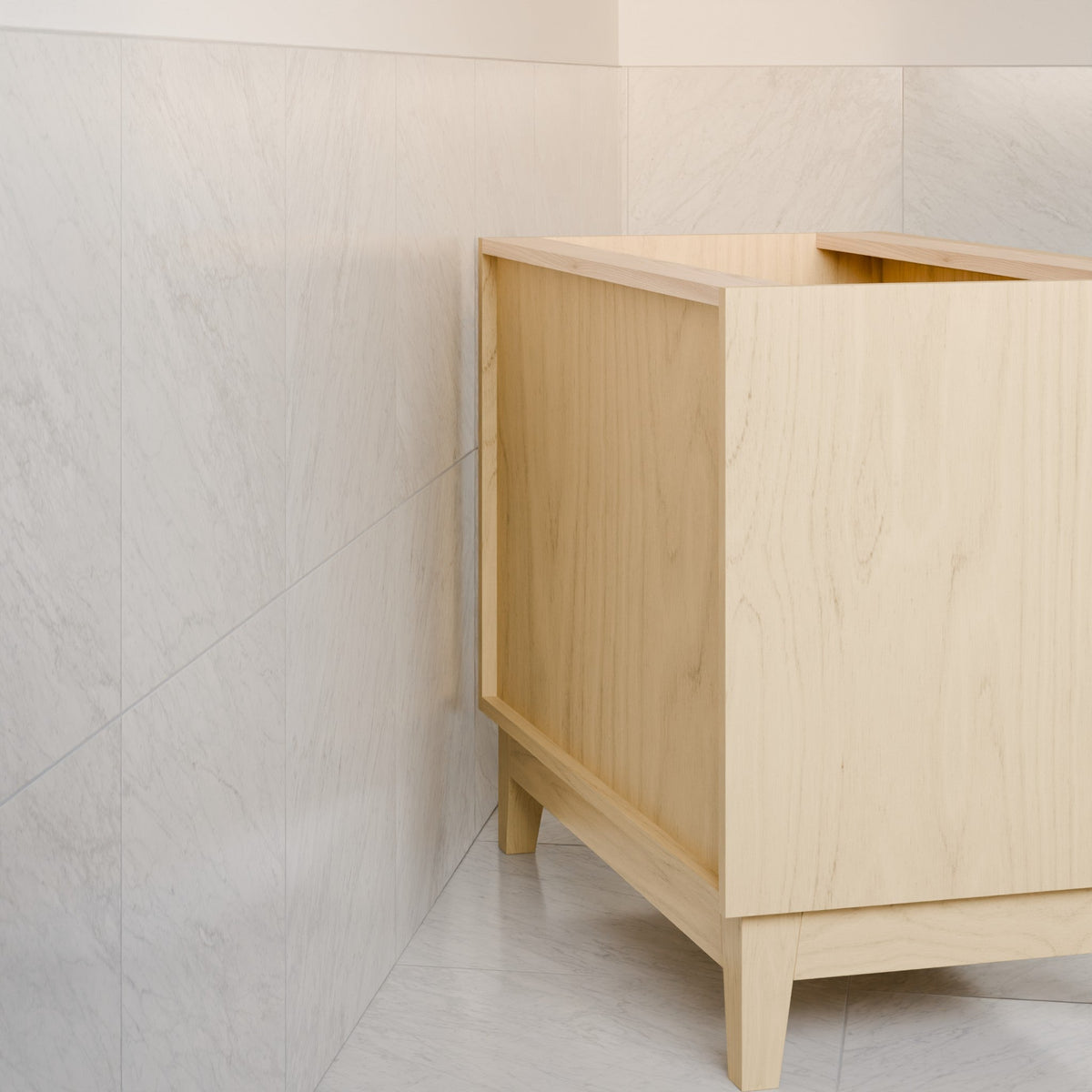 A light natural wood vanity cabinet with tapered legs, shown in a corner with light beige marble-look tiled walls and floor.