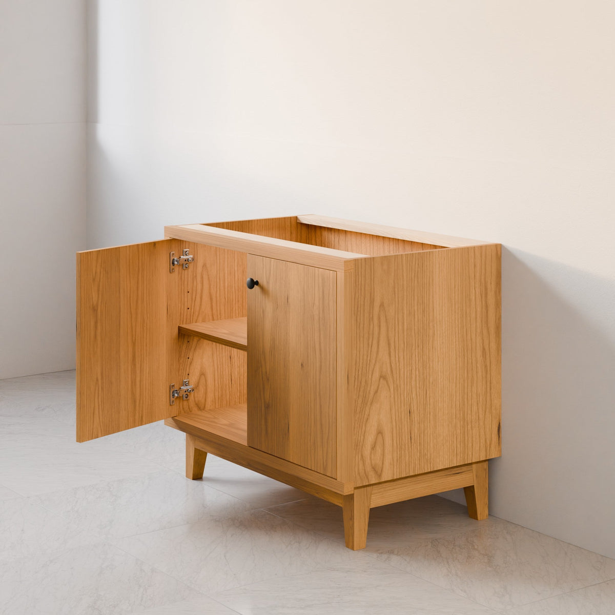 A light natural wood side cabinet with two doors, one open to reveal an interior shelf, set against a minimalist off-white wall and floor.